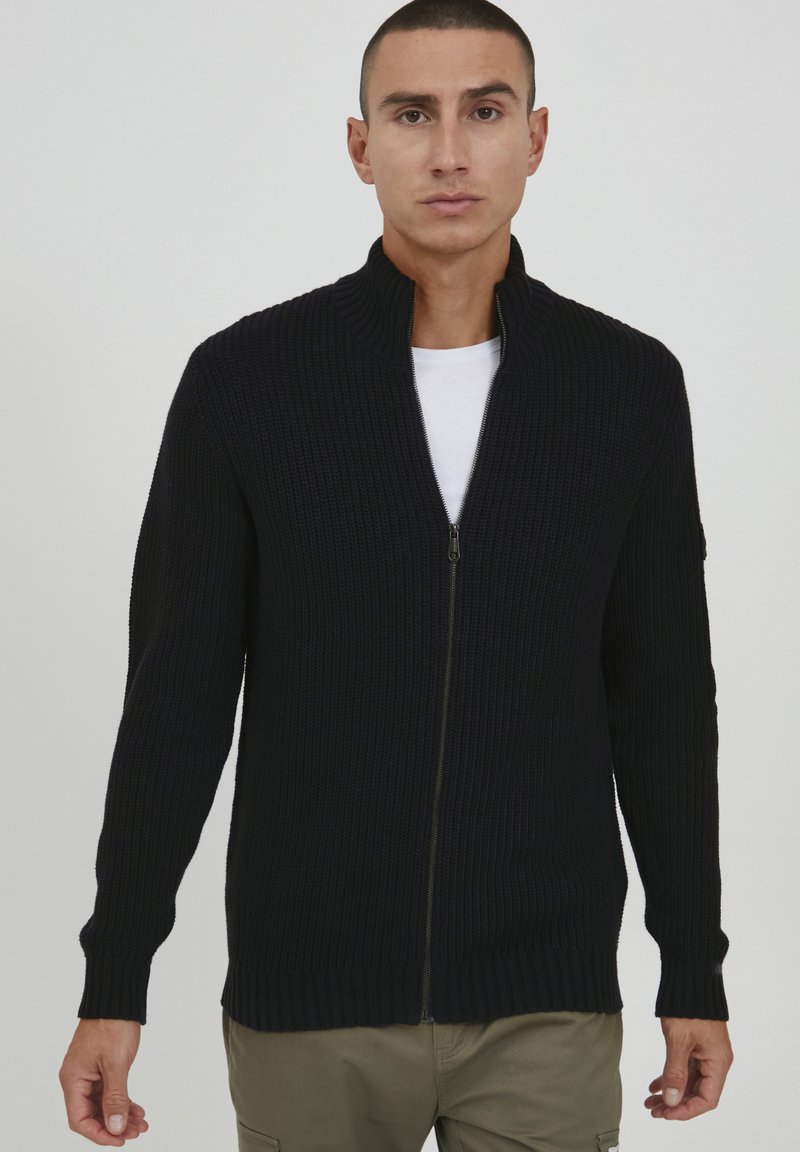 Maglione zip-up blu scuro con colletto alto, texture a coste, maniche lunghe e tasche laterali. Indossato sopra una camicia bianca. Finitura in tessuto liscia.