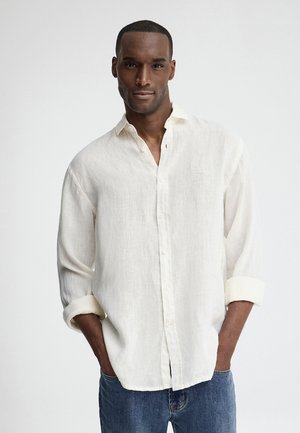 Man draagt een wit linnen overhemd met knopen en opgerolde mouwen en een blauwe spijkerbroek, staand tegen een effen lichte achtergrond.