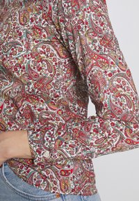 Chemise à manches longues avec des motifs colorés paisley et floraux en rouge, jaune et turquoise, portée avec un jean bleu clair sur fond blanc.