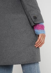 Šedý vlněný kabát s jedním černým knoflíkem, s barevným pleteným manžetem v růžové a modré, na pozadí světlých džínů.
