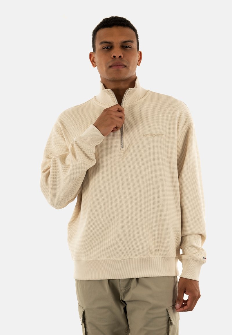 Sweatshirt beige zippé en tissu texturé, avec des poignets côtelés et un petit logo sur la poitrine. Le modèle porte un pantalon cargo clair.
