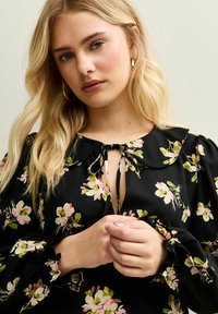 Robe noire à motifs floraux avec un col à nouer, des manches bouffantes et des poignets volants, ornée de motifs floraux roses et verts sur un tissu soyeux.