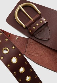 Καφέ δερμάτινη ζώνη με αγκράφα από χρυσαφί μέταλλο, διακοσμημένη με τρούκ και κυκλικούς κρίκους στη λουρίδα, με υφή φινιρίσματος.