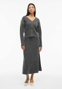 Grijze gebreide cardigan met een V-hals en knoopsluiting, gedragen over een bijpassende lange rok. Heeft een geribbelde structuur en lange mouwen.