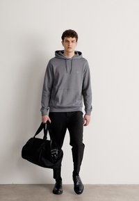Šedá mikina s kapucí a přední kapsou, spárovaná s černými úzkými kalhotami a černými botami. Drží velkou černou sportovní tašku. Minimalistický design.
