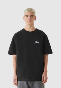 Črna bombažna majica s kratkimi rokavi, sproščenega kroja, z svetlo vijolično logotipom na levi strani prsi. Končani robovi z običajnim okroglim izrezom.