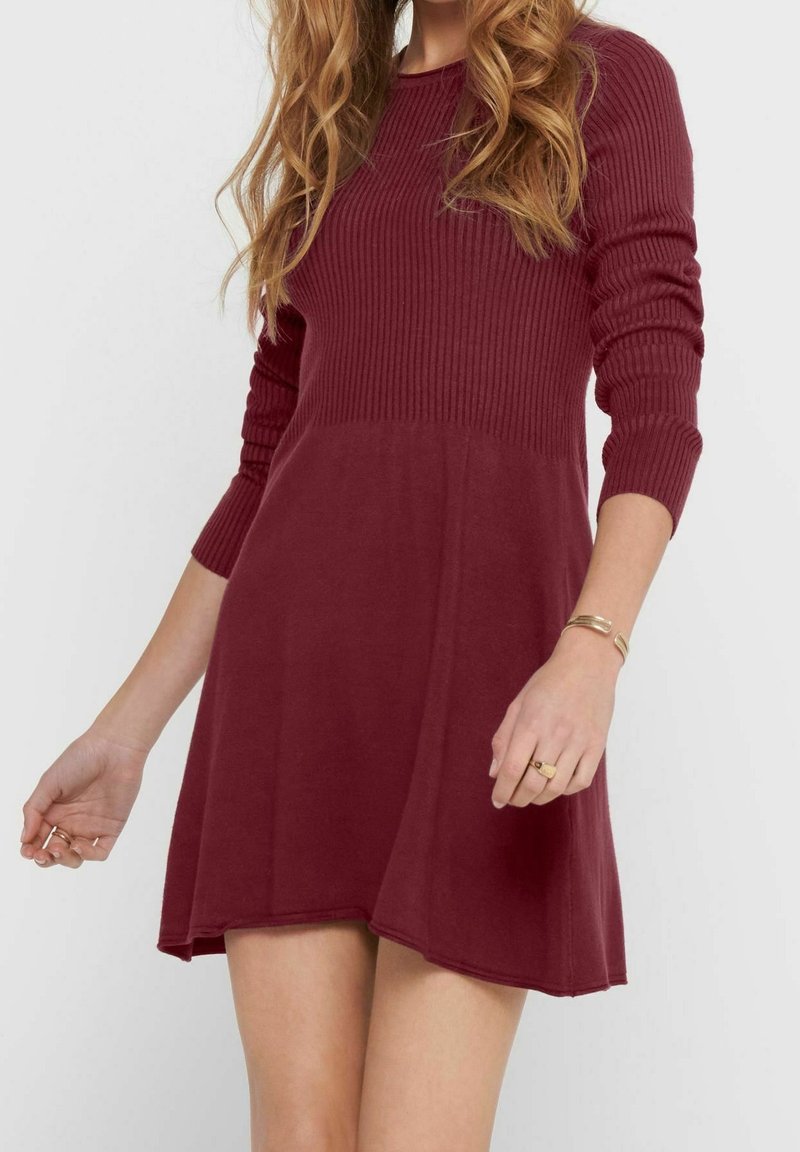Femme portant une robe bordeaux en maille côtelée à manches longues avec une jupe évasée, un bracelet en or et des bagues à la main gauche sur un fond uni.