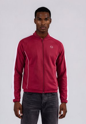 Homme portant une veste zippée grenat avec des bandes blanches sur les manches et un jean foncé, debout devant un fond uni clair.