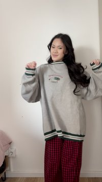 Mladý človek s dlhými tmavými vlasmi, oblečený v nadmerne veľkej sivej mikine a červených károvaných nohaviciach, gestikuluje zdvihnutými rukami proti jednoliatej bielej stene.