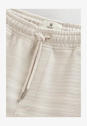 Pantaloni de trening bej deschis, cu un design texturat cu nervuri, având o talie ajustabilă cu șnur și un eticheta de marcă în partea de sus.