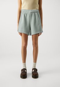 Pantaloni corti in tessuto morbido di un verde chiaro, con vita elastica, taglio dritto e tasche laterali, abbinati a calzature marroni e calze chiare.