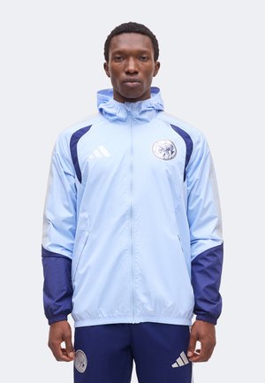Modèle masculin portant une veste Adidas bleu clair avec capuche et accents bleu marine, debout droit devant un fond clair uni.