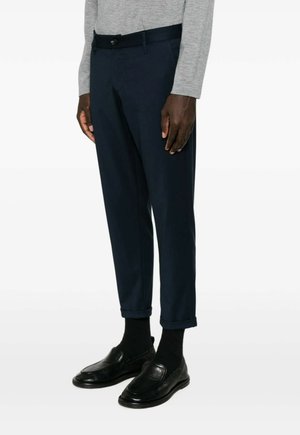 Man draagt marineblauwe cropped broek, zwarte sokken, zwarte leren loafers en een lichtgrijs shirt met lange mouwen, staand tegen een witte achtergrond.