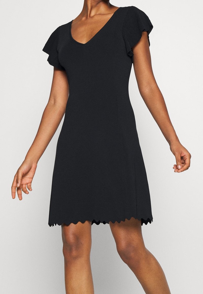 Schwarzes Kleid mit fließenden Ärmeln, V-Ausschnitt und gewelltem Saum. Der Stoff wirkt glatt und sorgt für eine figurbetonte Silhouette und Eleganz.
