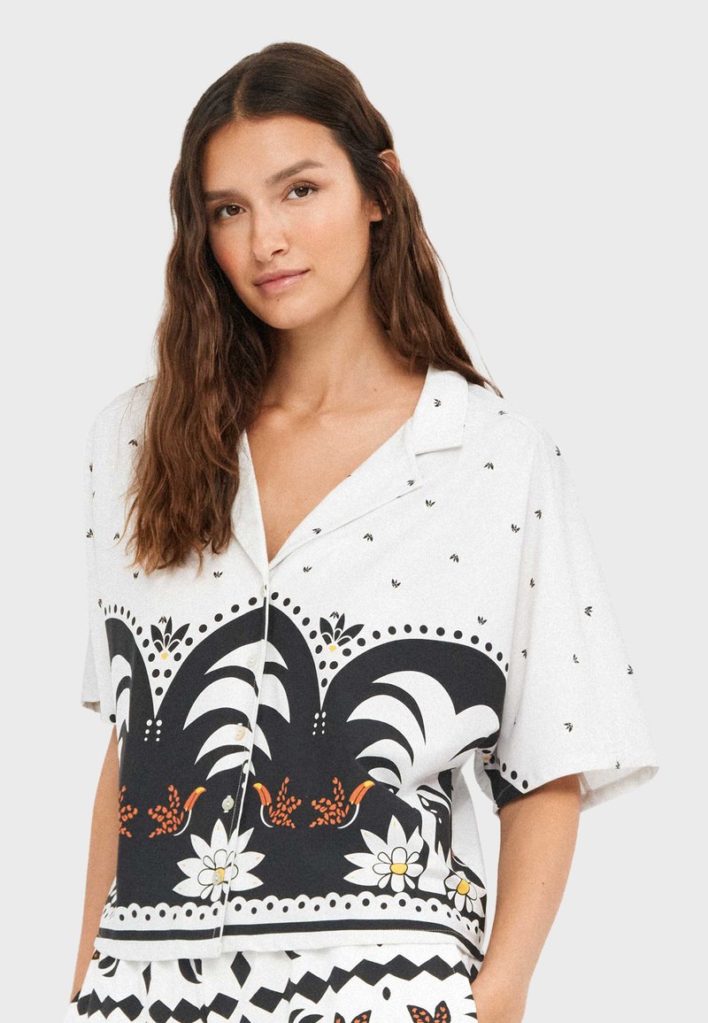 Femme aux longs cheveux bruns portant une chemise blanche à manches courtes avec un motif floral et d'oiseaux noir et orange.