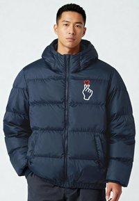 Giacca imbottita blu navy con cappuccio, caratterizzata da sezioni trapuntate, zip frontale e una grafica di una mano con un cuore rosso. Materiale morbido e lucido.