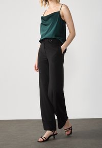 Camicetta verde in satin con spalline sottili, abbinata a pantaloni neri a vita alta e gamba larga con tasche frontali e una cintura nera. Sandali neri con cinturini.