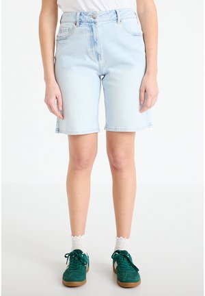Lichtblauwe denim shorts met hoge taille gedragen door een staande persoon, gecombineerd met witte sokken en groene sneakers op een witte achtergrond.