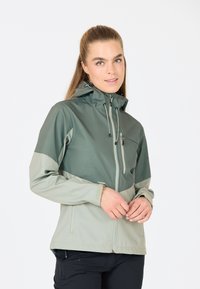 Mjukskaljacka i gröna nyanser, med dragkedja, justerbar huva och elastiska ärmslut. Materialet verkar lätt och har en mjuk textur.