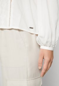 Camicia bianca con maniche arricciate, caratterizzata da un delicato inserto in pizzo e un'etichetta metallica con marca. Abbinata a pantaloni beige con tasche laterali.