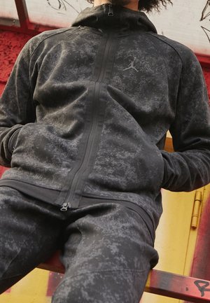Person trägt einen schwarz-grau gemusterten Kapuzenpullover mit Reißverschluss und passende Hose, sitzt auf einer rot-gelb mit Graffiti besprühten Bank.