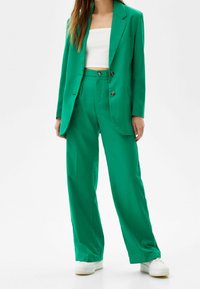 Veston vert accompagné d'un pantalon large assorti en tissu lisse. Le veston comporte deux boutons et le pantalon a une coupe décontractée. Top blanc en dessous.