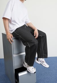 Pantalones de mezclilla negros con costuras blancas, camiseta blanca de gran tamaño y zapatillas blancas con acentos rojos, sentado en un archivador plateado.