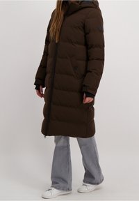 Brun puffjacka med horisontellt quiltat mönster, hel dragkedja, huva och sidofickor. Matchad med ljusgrå utsvängda jeans och vita sneakers.