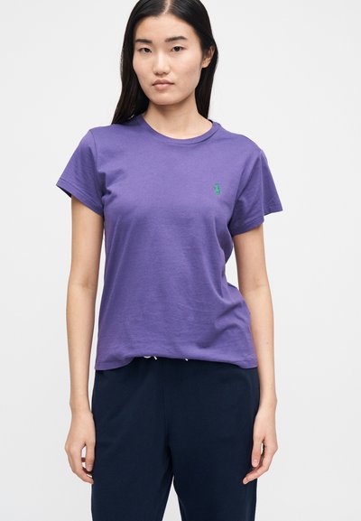 Lila pamutpóló kerek nyakkivágással és rövid ujjakkal, a bal mellrészen kis zöld logóval. Sima textúra, kényelmes szabás.