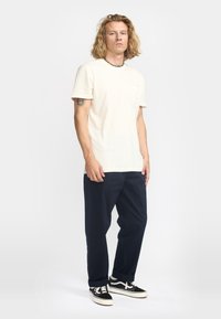 Cremfarbendes T-Shirt mit kurzen Ärmeln und Brusttasche, kombiniert mit dunkelblauen Hosen und schwarzen Sneakers mit weißen Akzenten und Schnürsenkeln.