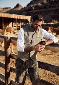 Hombre vestido con ropa tradicional bávara se apoya en una cerca de madera, mirando su reloj de pulsera, con vacas y montañas al fondo.