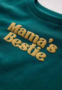 Tealfärgat tyg med gul upphöjd text som lyder "Mama's Bestie" på en texturerad sweatshirtkrage synlig på toppen.