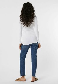 Langärmliges weißes Oberteil kombiniert mit blauen Jeans mit ausgefransten Säumen. Das Model steht mit lockigem Haar und zeigt eine schlichte, lässige Kleidung.