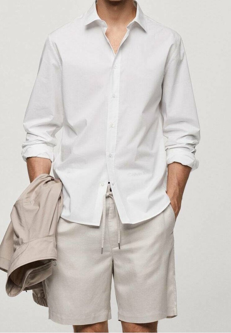 Biała koszula z guzikami i podwiniętymi rękawami, w połączeniu z jasnobeżowymi szortami. Beżowa kurtka jest zarzucona na lewym ramieniu.