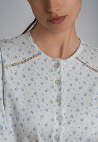 Maglietta button-up floreale blu chiaro con scollatura rotonda, dettagli in pizzo sulle spalle e cinque bottoni coordinati sul davanti.