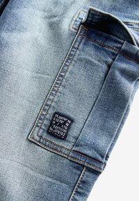 Lichtblauwe denimstof met een gestikt rechthoekig zakje en een klein zwart label met geografische coördinaten en editiegegevens.