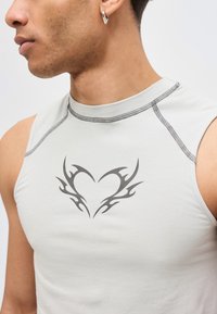 Κοντινό πλάνο ενός άνδρα που φοράει λευκό αμάνικο τοπ με μαύρο φυλετικό σχέδιο καρδιάς και ασημένιο κρίκο σκουλαρίκι στο αριστερό αυτί του.