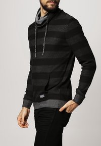 Czarny i szary pulower w paski z golfem, sznurkami i ściągaczem na dole. Miękki materiał, kieszeń z przodu oraz dopasowane rękawy.