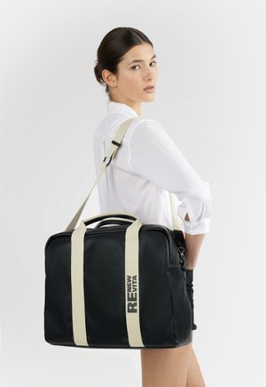 Donna con camicia bianca che porta una borsa rettangolare nera con manici beige e testo audace "RE NEW EVITA" sulla spalla, che guarda indietro.