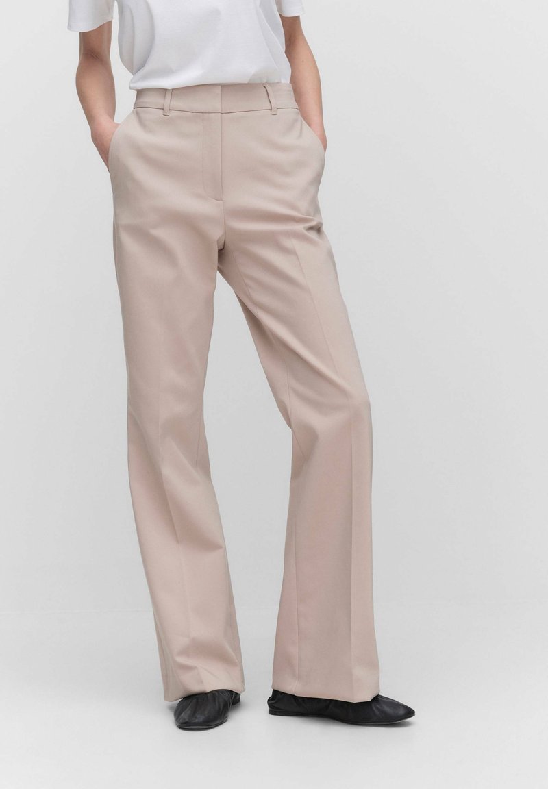 Personne portant un pantalon beige droit avec les mains dans les poches, associé à un haut blanc et des chaussures plates noires sur un fond uni.