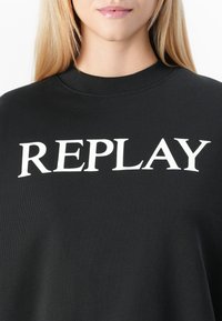 Blonde persoon die een zwart sweatshirt met ronde hals draagt, met grote witte letters "REPLAY" op de borst.