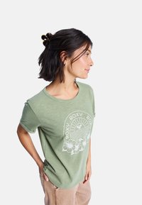 T-shirt vert à manches courtes avec un graphique floral blanc et le texte "ROXY". Fabriqué en matériau léger, il présente une coupe décontractée et un col rond.