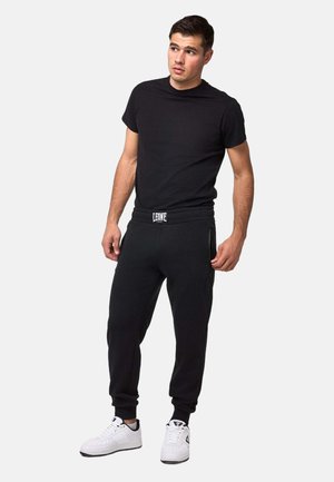 Jeune homme portant un t-shirt noir, un pantalon de jogging noir avec le logo "Leone" et des baskets blanches, debout devant un fond clair uni.
