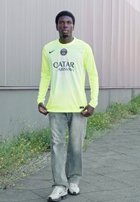 Maillot de sport à manches longues jaune néon avec des accents noirs et le logo de Qatar Airways, associé à un jean bleu clair et des baskets blanches.