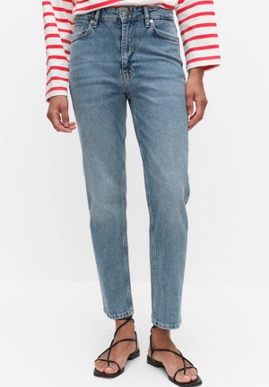 Osoba ubrana w jasnoniebieskie jeansy z wysokim stanem, czerwonobiałą pasiastą bluzkę z długim rękawem oraz czarne, płaskie sandały na paski na białym tle.