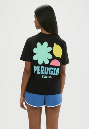 Frau mit langen schwarzen Haaren trägt ein schwarzes T-Shirt mit bunten grafischen Formen und dem Schriftzug "PERUGIA Ellesse" sowie blaue Shorts.
