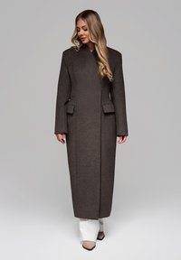 Lang brun uldfrakke med lige snit, brede ærmer og forlommer, sammen med hvide bukser med bred ben og beige spidse sko.