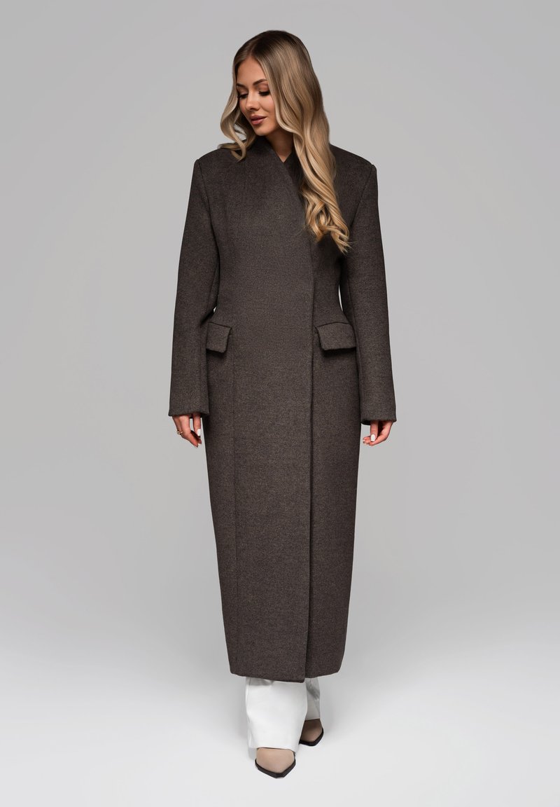 Lang brun uldfrakke med lige snit, brede ærmer og forlommer, sammen med hvide bukser med bred ben og beige spidse sko.