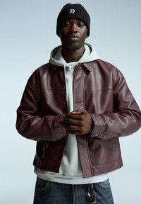 Veste en cuir bourgogne avec un design à col, portée sur un hoodie gris. La personne porte également un bonnet en tricot noir et un jean en denim.