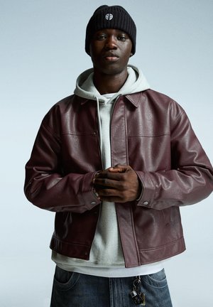 Burgundy leather jacket with a collared design, worn over a gray hoodie. The person is also wearing a black knit beanie and denim jeans.